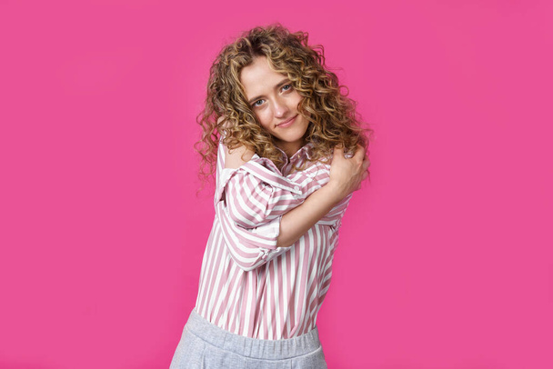 Waiting for a special moment. Portrait of a smiling woman who hugs herself with her arms. Isolated on a pink background. People sincere emotions concept. - Foto, afbeelding