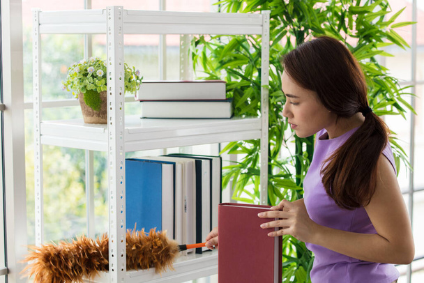 Aziatisch jong vrouw of meid is vegen schoonmaken stof op de boekenplank met een veer stofdoek thuis. - Foto, afbeelding