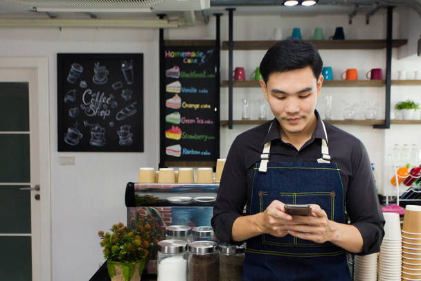 The coffee shop entrepreneur is checking customer orders on the cell phone while standing in front of the counter. Asian young barista wearing the apron in the cafe. - Photo, Image