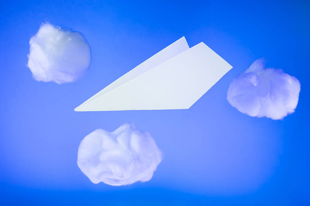 Белый бумажный самолет пролетает среди облаков ваты на синем фоне. Креативная концепция путешествий, перелетов. - Фото, изображение