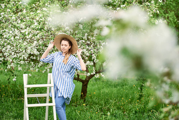 Çiçekli bir bahar bahçesinde kokunun tadını çıkaran güzel, mutlu genç bir kadın. Beyaz merdivenlerin yanındaki bahçede şapkalı genç bir kadın duruyor. - Fotoğraf, Görsel