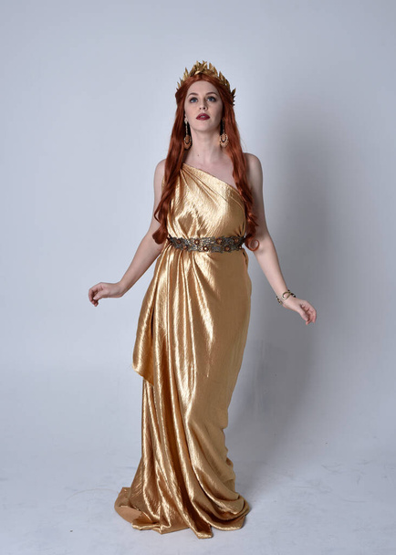 Portrait complet de fille aux cheveux roux portant une longue toge grecque et une couronne dorée. pose debout isolée sur un fond de studio gris. - Photo, image