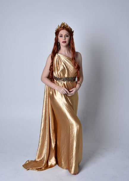 Retrato de cuerpo entero de niña de pelo rojo con toga larga y corona dorada. De pie posan aislado contra un fondo gris estudio. - Foto, imagen