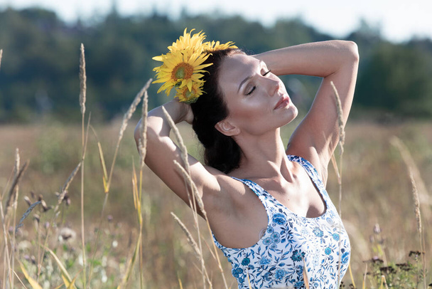 brünette Frau auf dem Feld, Sonnenblume im Haar, sonniger Tag, sie ist in einer Sommersonne. - Foto, Bild