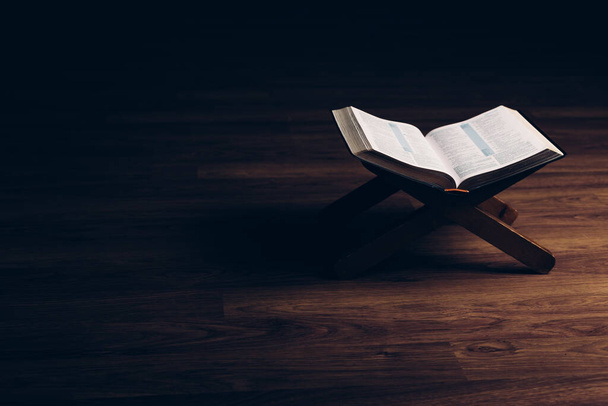 відкрита Біблія на книжковій підставці на чорному тлі з дерев'яною підлогою і м'яким світлом
 - Фото, зображення
