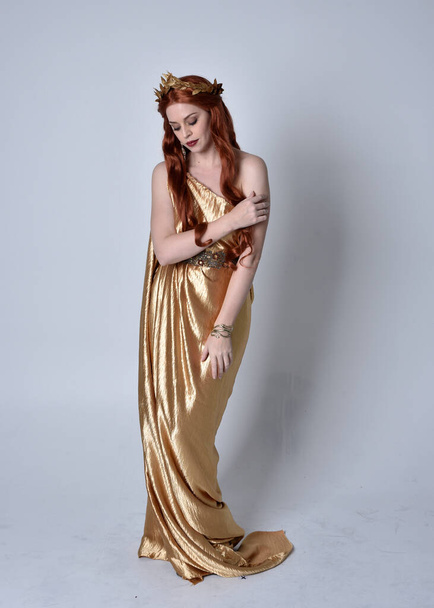 Full length portrait of girl with red hair wearing long grecian toga and golden wreath. Standing pose iisolated against a grey studio background. - Фото, изображение
