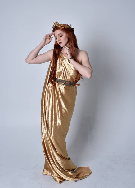 Full length portrait of girl with red hair wearing long grecian toga and golden wreath. Standing pose iisolated against a grey studio background. - Photo, Image