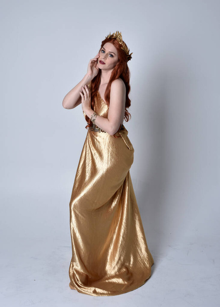 Full length portrait of girl with red hair wearing long grecian toga and golden wreath. Standing pose iisolated against a grey studio background. - Foto, immagini