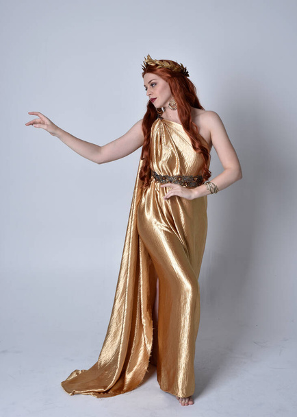Full length portrait of girl with red hair wearing long grecian toga and golden wreath. Standing pose iisolated against a grey studio background. - Photo, Image