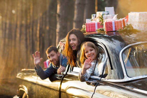 Mère heureuse père et petite fille regardant par la fenêtre de la voiture avec des cadeaux de Noël sur le toit et saluant les mains - Photo, image