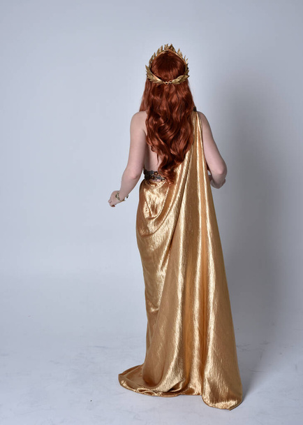 Full length portrait of girl with red hair wearing long grecian toga and golden wreath. Standing pose with back to the camera,  isolated against a grey studio background. - Photo, Image