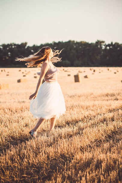 Happy young woman in dress walking in evening in field with hay bales, beautiful romantic girl with long hair outdoors in field - Φωτογραφία, εικόνα