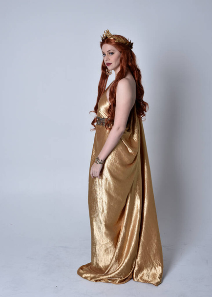 Pełna długość portret dziewczyny z czerwonymi włosami w długich greckich toga i złoty wieniec. Pozycja stojąca tyłem do kamery, odizolowana na szarym tle studia. - Zdjęcie, obraz