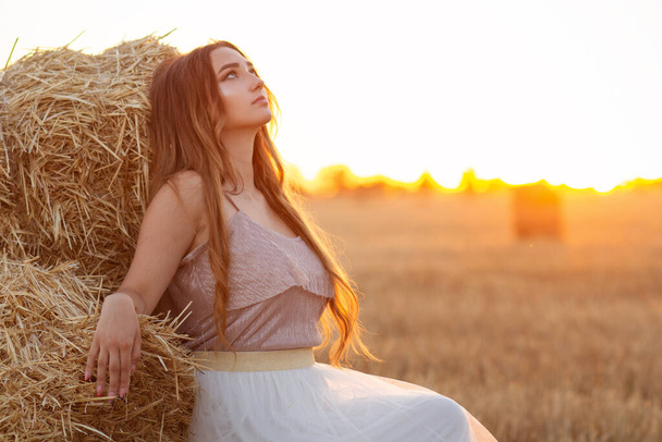 Happy woman sitting on hay stack walking in summer evening, beautiful romantic girl with long hair outdoors in field at sunset - Foto, immagini