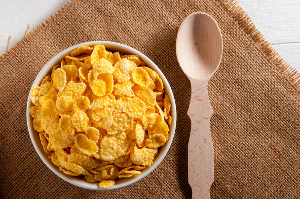 White bowl of cornflakes, next to a wooden spoon. Everything is on a sackcloth napkin on a white wooden table. Horizontal frame. - Foto, immagini