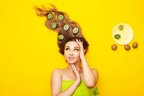 fashion portrait of beautiful girl lying on colored background with kiwi fruit slices spread out on long hair, people and food, young peaceful woman takes care of beauty and body - Foto, afbeelding