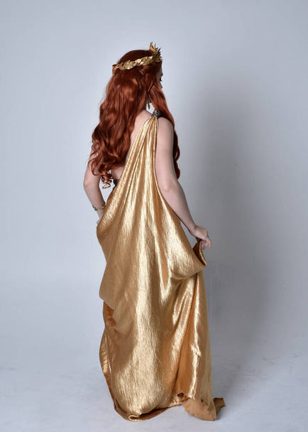 Ολόσωμο πορτραίτο κοριτσιού με κόκκινα μαλλιά που φορούσε μακρυμάνικη τήβεννο και χρυσό στεφάνι. Μόνιμη θέτουν με την πλάτη στην κάμερα, απομονώνονται σε ένα γκρι φόντο στούντιο. - Φωτογραφία, εικόνα
