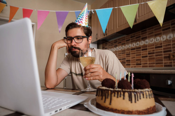 Αναστατωμένος νεαρός που κάθεται στην τούρτα γενεθλίων και κοιτάζει με θλιμμένα μάτια. Έννοια της μοναξιάς σε καραντίνα κατά τη διάρκεια της Πανδημίας του Coronavirus COVID-19. - Φωτογραφία, εικόνα