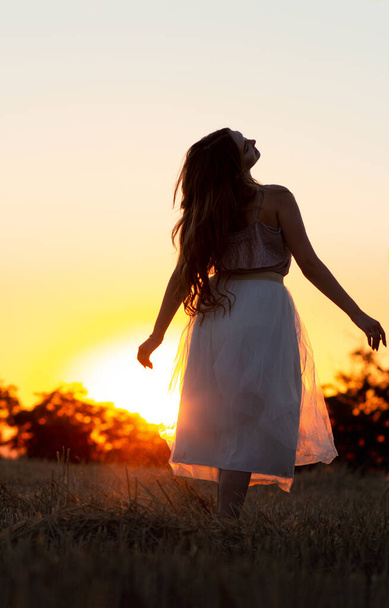 σιλουέτα φιγούρα του όμορφου ονειρεμένου κοριτσιού με φόρεμα στο ηλιοβασίλεμα σε ένα πεδίο, νεαρή γυναίκα με μακριά μαλλιά απολαμβάνοντας τη φύση στον ορίζοντα - Φωτογραφία, εικόνα