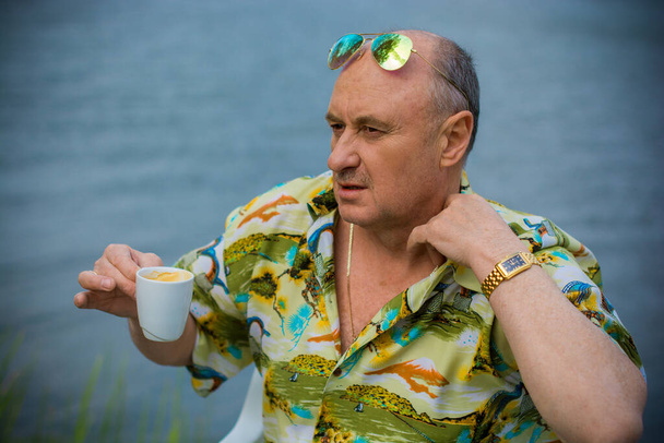 Een man van volwassen leeftijd, Amerikaans uiterlijk, rustend bij een blauw meer in een Hawaïaans shirt, strohoed en groene bril, een vrolijk positief en emotioneel persoon - Foto, afbeelding