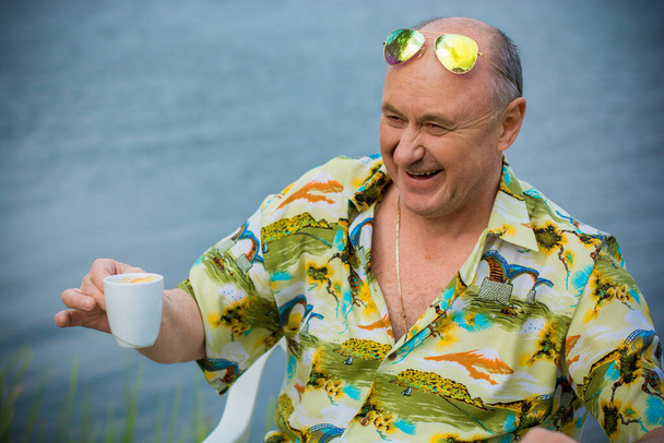 Muž zralého věku, amerického vzhledu, odpočívající u modrého jezera v havajské košili, slamáku a zelených brýlích, veselá pozitivní a emocionální osoba - Fotografie, Obrázek