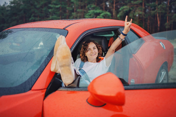 Φθινόπωρο οδικό ταξίδι, ελευθερία και σόλο ταξιδιώτη. Όμορφη γυναίκα κάθεται στο αυτοκίνητο στην όχθη της θάλασσας, τα πόδια στο παράθυρο του αυτοκινήτου, κοιτάζοντας το όμορφο ηλιοβασίλεμα μετά τη βροχή, πέφτει στο παρμπρίζ, πτώση υπαίθριο τρόπο ζωής - Φωτογραφία, εικόνα