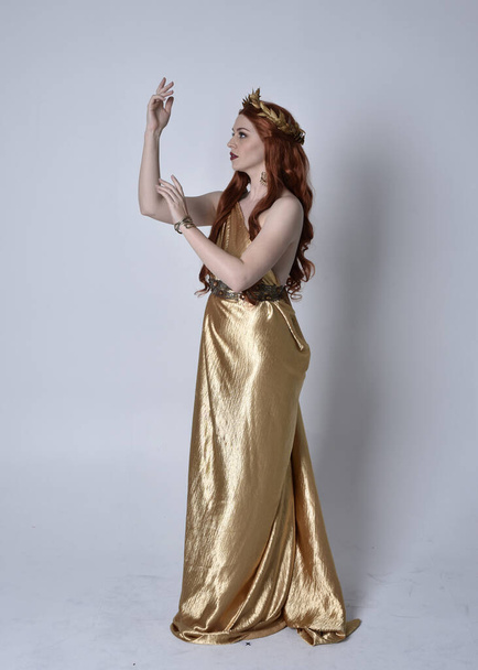 Full length portrait of girl with red hair wearing long grecian toga and golden wreath. Standing pose in side profile,  isolated against a grey studio background. - Photo, Image