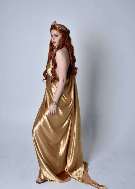 Портрет девушки с рыжими волосами в длинных греческих тогах и золотом венке. Стоячая поза в боковом профиле, изолированная на сером фоне студии. - Фото, изображение