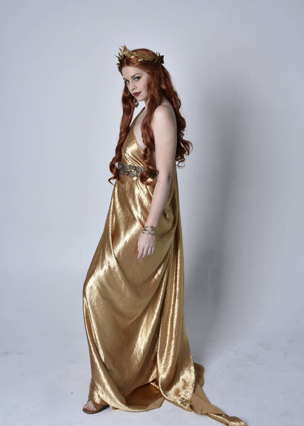 Portrait complet de fille aux cheveux roux portant une longue toge grecque et une couronne dorée. Pose debout de profil latéral, isolée sur fond de studio gris. - Photo, image