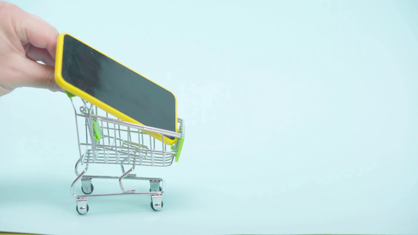 Vista cortada do homem montando carrinho de compras com smartphone no fundo azul - Filmagem, Vídeo