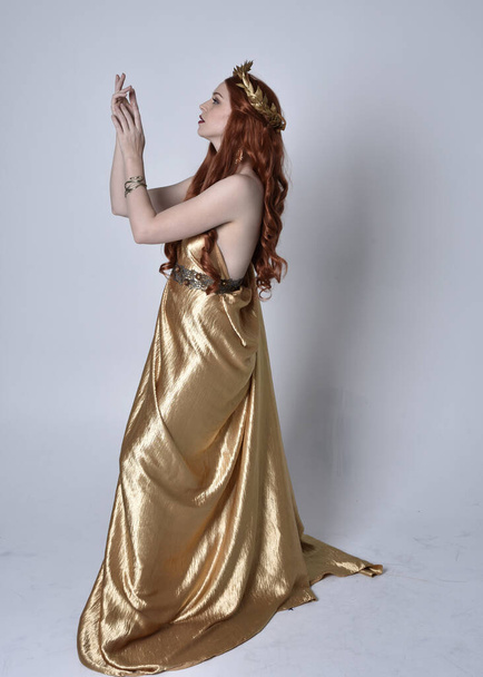 Full length portrait of girl with red hair wearing long grecian toga and golden wreath. Standing pose in side profile,  isolated against a grey studio background. - Photo, Image