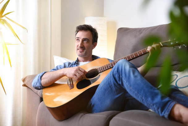 Relaxed and smiling man lying on a sofa playing the guitar in a room at home - Foto, immagini