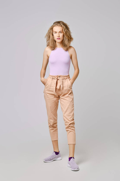 Pretty girl with blonde messy hair in tank top and trousers stands with hands in pockets - Foto, Imagen