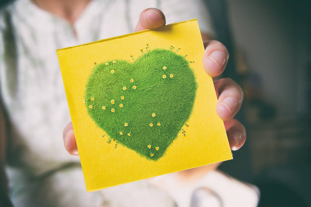 Woman's hands holding sticky note with green heart with yellow flowers, natural and ecological life style concept, gardening in pure, healthy environment - Photo, Image