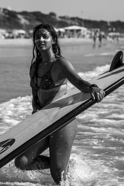Pystysuora harmaasävykuva valkoihoisesta naisesta, jolla on aurinkorasva kasvoillaan ja surffilauta rannalla Cádizissa, Andalusiassa, Espanjassa - Valokuva, kuva