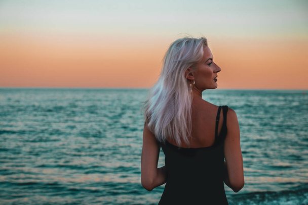 Jeune belle femme blonde sur la plage pendant le coucher du soleil regardant droit à l'horizon - Photo, image