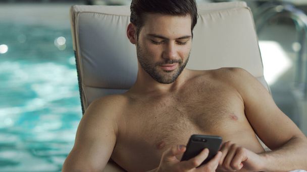 Closeup man looking phone by pool. Male relaxing with phone at wellness center - 写真・画像