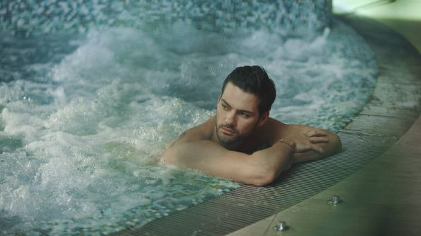 Σέξι τύπος χαλαρώνει στην πισίνα στο ξενοδοχείο σπα. Ο άνθρωπος αγγίζει βρεγμένα μαλλιά στο τζακούζι στο spa - Φωτογραφία, εικόνα