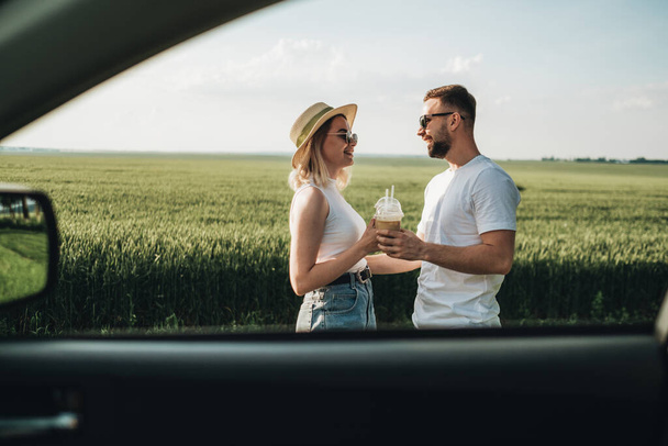 Hombre y mujer pasando un buen rato cerca del coche, tomando café frío y disfrutando de un viaje por carretera de aventura - Foto, imagen