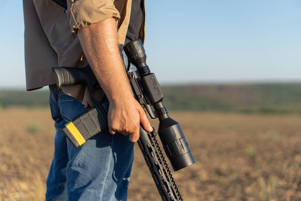 Un rifle en la mano de un hombre, de cerca. Cazador con un arma en la caza contra el fondo de un hermoso paisaje de verano - Foto, imagen