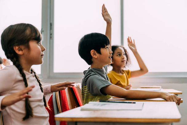 Elementary age Asian student boy raised hands up in Q and A classroom. Diverse group of pre-school pupils in elementary age in education building school. - Zdjęcie, obraz