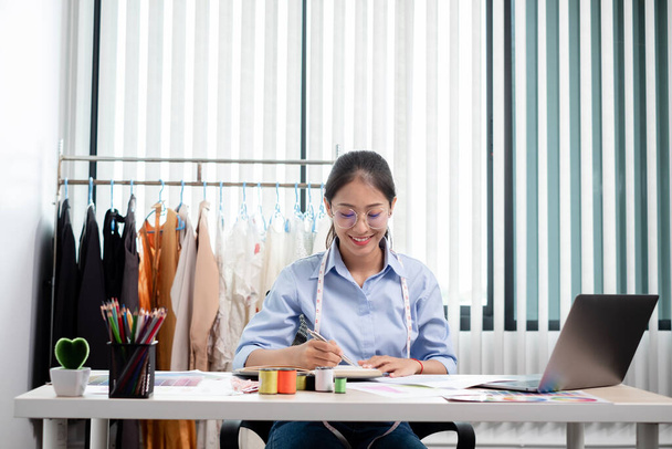 Schöne junge asiatische Modeschneiderin mit Textilnähaccessoires und Entrepreneurskizzen sind voller heller Farben auf dem Schreibtisch mit einem Laptop für kreative Ideen im Atelier. - Foto, Bild