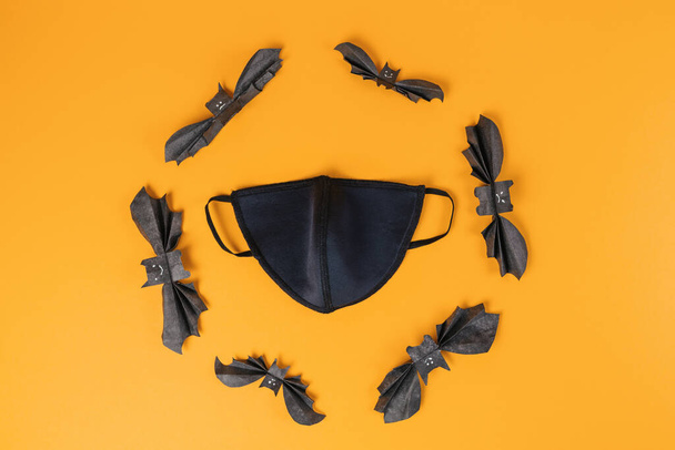 Masque anti-virus noir réutilisable entouré de chauves-souris en papier. Fond orange. Pose plate. Espace de copie. Le concept de protection contre le virus pendant Halloween. - Photo, image