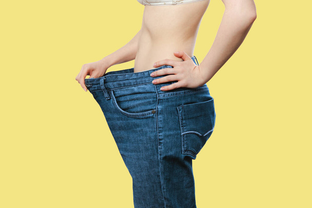 Una donna caucasica mostra i risultati della sua perdita di peso tirando su jeans blu a vita larga. Isolato, sfondo giallo. Vista laterale. Il concetto di perdita di peso, sport e dieta. - Foto, immagini