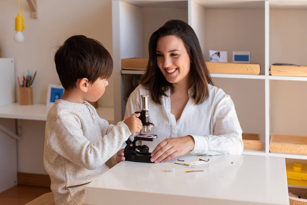 Lillte Kid explorando la ciencia con un microscopio y la ayuda de la madre o el maestro. Homeshooling. Comunidad de aprendizaje. Escuela Montessori - Foto, Imagen