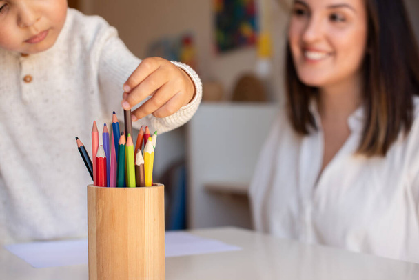 Ένα μικρό παιδί προσχολικής ηλικίας ζωγραφίζει με χρωματιστά μολύβια με μητέρα ή εκπαιδευτή δασκάλων. Συγκεντρώσου στα μολύβια. Μαθήματα κατ 'οίκον. Μαθαίνοντας κοινότητα. Σχολή Μοντεσόρι. - Φωτογραφία, εικόνα
