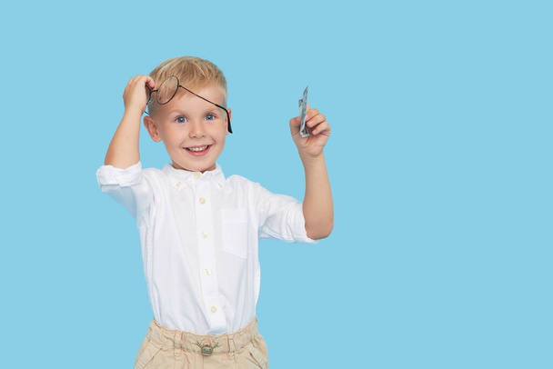 Smart child boy in glasses and a white shirt holds a paper dollar bill on a blue background. The concept of beneficial loans for childrens education. Wow face - Фото, зображення