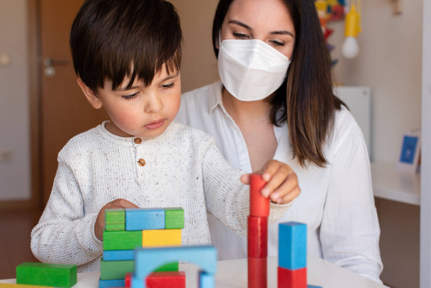 Liltle Preescolar Kid jugando con bloques de madera y educador maestro ayudan a usar la máscara facial para la pandemia de coronavirus. Homeshooling. Comunidad de aprendizaje. Escuela Montessori - Foto, Imagen