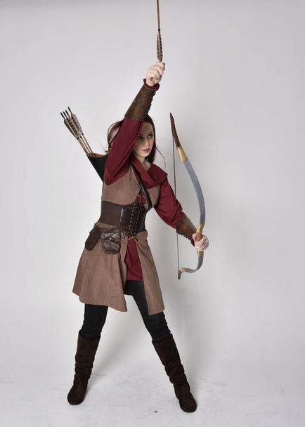 Retrato de longitud completa de chica con el pelo rojo con traje de arquero medieval marrón.. Posar de pie sosteniendo un arco y flecha, aislado sobre un fondo gris estudio. - Foto, imagen