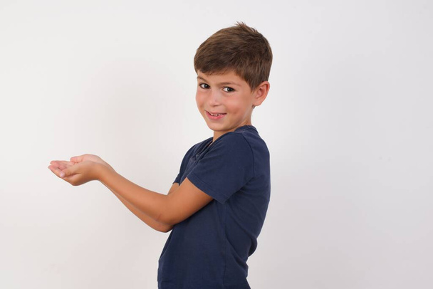 Krásný chlapec nosí ležérní tričko stojící nad izolovaným bílým pozadím, ukazuje stranou s rukama otevřenýma dlaněmi ukazujícíma kopírovací prostor, prezentuje reklamu s nadšeným úsměvem - Fotografie, Obrázek